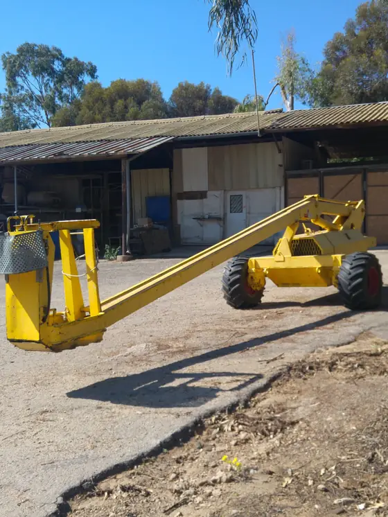 A yellow self-propelled cherry picker parked in a farmyard, its single boom lowered with the one-person picking cage near the ground